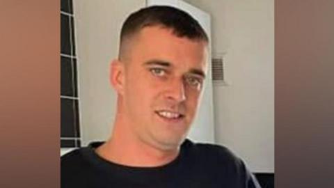Aidan Matthews is smiling and wearing a dark top in a kitchen with tiles and cupboards behind him.
