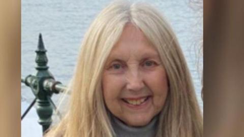 Ellen Cook is pictured smiling at the camera and wearing a grey jumper. She has long blond and white hair