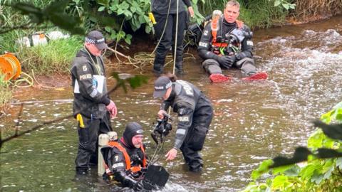 Divers in the River Irwell - a man in a police hat stands up - with a man sitting in the water beneath him with oxygen tanks on his back - a woman in a police hat and outfit holds a camera