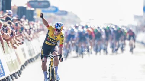 Cyclist Wout van Aert is pictured crossing the final stage finish line of the Tour of Britain in 2023, which he won. He is wearing a yellow and black cycling suit while riding his yellow bike.