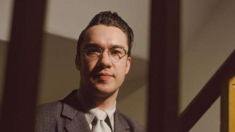 Mark Lamarr in glasses, a quiff, suit and tie in an official portrait photo taken in 1995