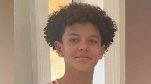 Noah smiling at the camera. He is wearing a white football shirt with red edging and has short curly hair.