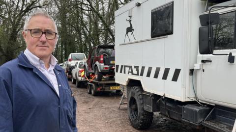 A man with grey hair and black framed glasses is standing looking at the camera. He is wearing blue overalls. He is stood next to a fleet of 4x4 vehicles.
