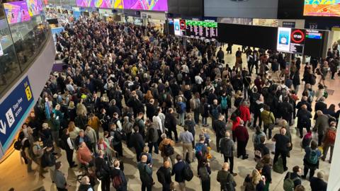 The concourse at London Waterloo pictured from above. It is full of passengers standing in queues and in groups, stretching away to the edge of the frame. Many are looking up at the electronic departures boards. 