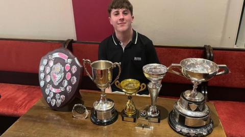 A boy in a black t-shirt, who is sitting down, with seven trophies.