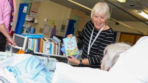 A smiling volunteer holding a book in front of an anonymous female patient with a trolley filled with books behind her