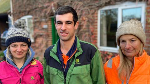 A man stands in the middle of two women. He is wearing a green farming overall and has dark short hair. The woman to his left is wearing a pink coat and wooly hat. The woman to his right is wearing an orange coat and cream wooly hat.