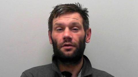 A custody picture of a man with short hair, facial hair, who wears a dark collared shirt.