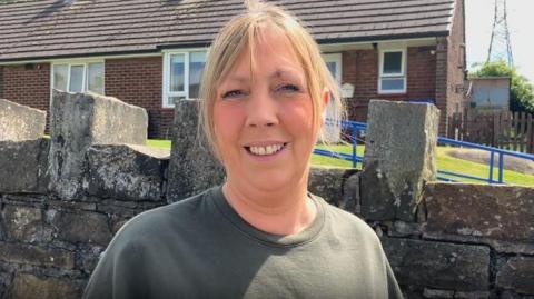 Michelle Boult with blond hair tied back wearing a dark green t-shirt standing in front of a stone garden wall with a house behind her. She is smiling.