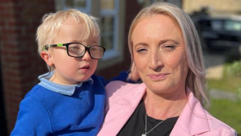 Head and shoulders shot of five-year-old Harvey Hind with blonde hair wearing black glasses and royal blue school sweatshirt over a pale blue polo shirt. He is being held by his mum in the garden of their house in the sunshine. She has long straight blonde hair and is smiling and wearing a pale pink blazer over a black tshirt