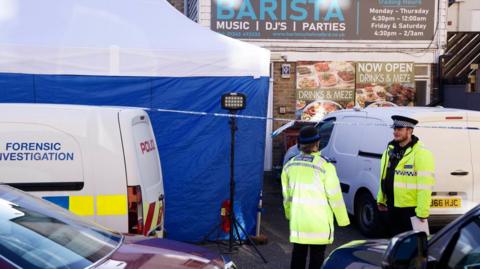 Two police officers in yellow high-vis jackets standing in front of a van parked in front of a food outlet called Barista. To its left is a large blue and white police forensic tent and in front of that is a white van with police and forensic investigation written on its sides 