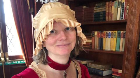 Mai Black smiles at the camera as she stands in front of a large wooden book case. She wears a gold coloured hat, red choker necklace and a red and gold dress. 
