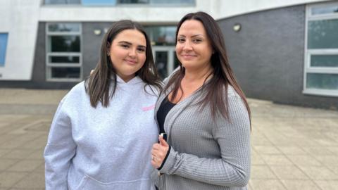 A mother and her teenage daughter smiling at the camera. Both are wearing grey and have long brown hair. 