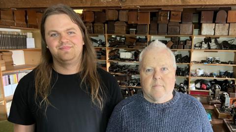 Gabriel has long hair and it standing next to his father with grey hair and hundreds of sewing machine paraphernalia in the background. 