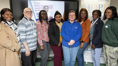 Seven women and one man posing for a photograph in a line. All those pictured are smiling at the camera. They are standing in a room with a green carpet. A pull-up banner saying HOPE is behind them to the right. On the left behind them is a digital screen with an image of a smiling young women and words partially obscured, but Your NHS can be made out.