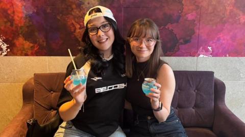 Caitlin and Katy sit on a purple sofa smiling while holding light blue BuzzBall canned cocktail drinks.