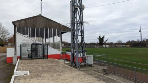 Part of the ground at Stag Meadow, with a covered terrace to the left of the picture and the grass pitch to the right, with the bottom part of a floodlight in the centre of the picture.