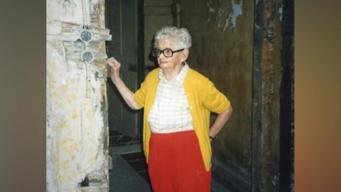 A woman stands in a rundown building wearing a bright yellow cardigan, white shirt and red trousers