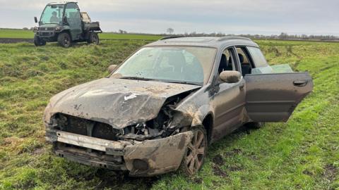 A dark-coloured car caked in mud, its front bumper hanging off, abandoned in a field in the Fens.
