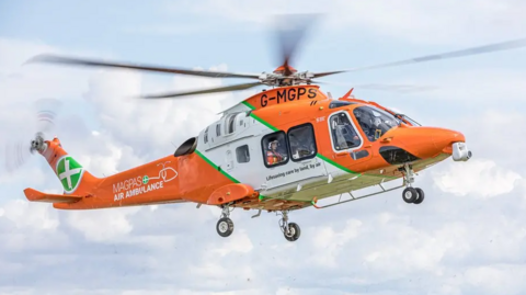 The Magpas helicopter taking off in flight. It is an orange and white aircraft with the logo emblazoned on the side and rotors whirring. An operative in a white helmet can be seen within the helicopter.