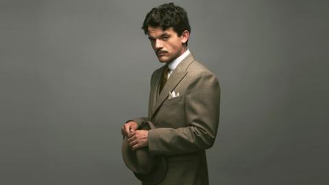 Edward Bluemel as Hercule Poirot, withh dark hair and a moustache, wearing a grey suit, holding a top hat