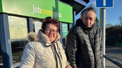Moira is wearing a white coat with a fluffy hood. She has short brown hair and is wearing glasses. Peter is wearing a blue puffa jacket and has short receding hair.