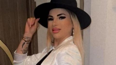 A woman, with blonde hair, wearing a black hat and white shirt. She is holding the hair with one hand. She has tattoos on this arm. She is smiling at the camera. 