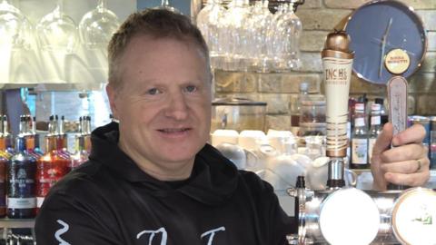 James stands behind the bar pulling a pint while facing the camera. In the background are gin bottles in different colours and stacked white tea cups. He is wearing a black hoodie with white writing on it saying the name of his pub, and has short dark hair.