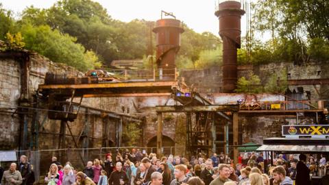 Crowds gathered on the Brymbo site in Wrexham. The framework of a steelworks can be seen above the crowd, along with green forestry behind the walls of the unit. 
