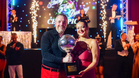 A man and woman stand in the middle of a dancefloor, holding a large glitterball trophy between them. Light from the trophy is being reflected onto the man's face