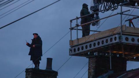 A man dressed in dark clothing stands on top of a chimney  in the early evening light. He is wearing a flat cap and holding his right arm above his head with a twig-like wand in his grasp. A member of film crew can be seen standing on a platform in front of him