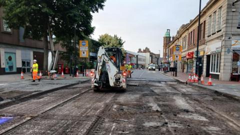A town centre road which is having new tarmac put down. It is covered with mud and tracks. A white vehicle is being driven along it while workers in yellow and orange hi-vis outfits move about the site. High street buildings and trees are visible on either side of the road.