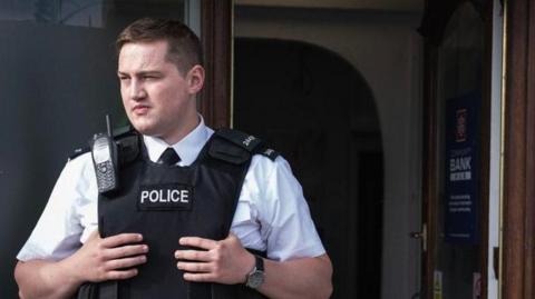 Finnian Garbutt, a man with dark hair, wearing a white shirt with black tie and a black support vest with the words police on it. He is wearing a black watch on his left wrist and there is a brown wooden door in the background.