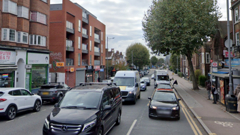 A Google Streetview image of Lea Bridge Road, with traffic, with shops on one side of the road and a bus stop on the other.