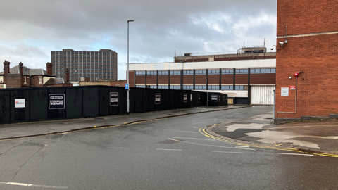 A road junction, with a brick building on the right hand side. Black hoarding surrounds a site on the left, with signs on them reading: Etruscan Square