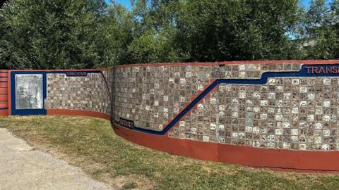 A long, meandering, decorative wall on a sunny day. Grass around it and trees behind. The wall has thousands of tiles each with an individual design. A blue line travels through the wall, bending, representing the river and splitting up the themes of the tiles.