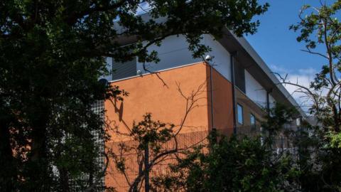 An exterior view of the Brook House detention centre. The photo is taken from outside the centre, so building itself is partly obscured by a high fence and the leaves of a tree.