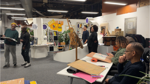 People gather in an art studio. Two men are looking at a book, while four people with easels watch