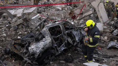 A firefighter uses water to clean his eyes as he stands next to the burned debris of a car destroyed in a Iranian strike on Israel