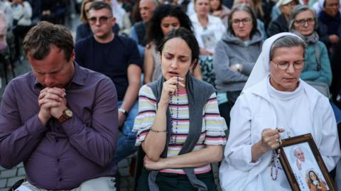 Three mourners side-by-side in prayer with crowds seated in rows behind them