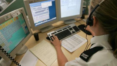 A woman with her hair pulled back into a ponytail sits at a desk with three screens and a keyboard. She has a headset on and is wearing a white blouse with a black epaulette labelled "ENQUIRY OFFICER" in white lettering.