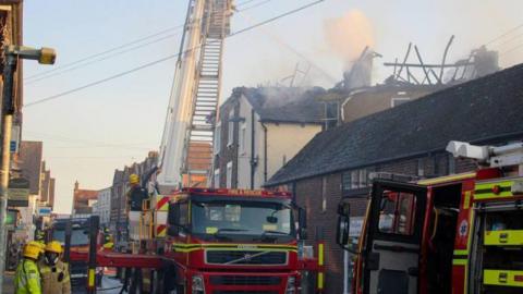 Fire engines and firefighters in a road of shops and flats above with smoke seen coming from the roof of a building.