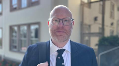 Deputy Rob Curgenven - A man with a bald head, wearing black rimmed glasses, with a ginger and grey beard. He is wearing a blue suit and a white colleared shirt, with a navy blue tie, with Guernsey flags on it. He his holding a navybag strap over his right shoulder. 