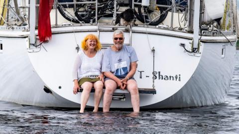 Debbie and Stephen sitting on the back of their yacht with their feet in the water. The word "T-Shanti" is printed on the back of the white boat. Both are smiling at the camera.