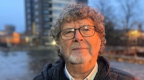 Mark Slater who is the Chairman of the Campaign to Save the Seven Sisters, stands in front of one of the Seven Sisters tower blocks on a wet day, with leaves scattered on the pavement around him. He is wearing black-rimmed glasses and has curly grey hair.