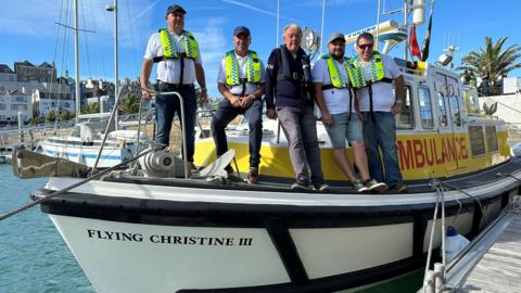 Five men are standing aboard Flying Christine III which is tied up on a pontoon in the harbour. They are wearing lifejackets and are all smiling. Behind them is a yacht and some houses overlooking the foreshore. 