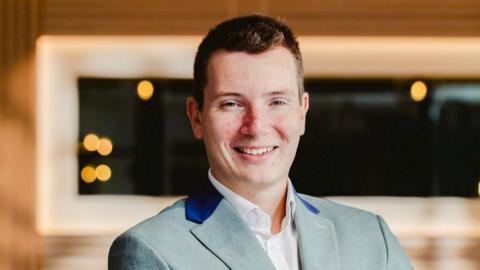A man with dark hair smiles into the camera. He is wearing a green suit jacket with flashes of blue on the lapels and a white shirt - unbuttoned at the neck - underneath.