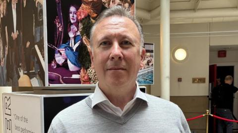 LIPA principal and chief executive Professor Sean McNamara has grey hair and wears a white shirt with a grey v-neck jumper. He stands in the LIPA building in front of a sign.