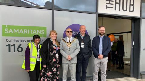 Five people, including Kirsten Rose and the Wolverhampton mayor, stand outside the glass frontage of The Hub at the station. One woman works for WMR and has a florescent bib over her clothing. The mayor is wearing a suit with his mayoral chains on top with two other men in navy and grey casual clothing. Kirsten is wearing a floral dress and dark jacket.
