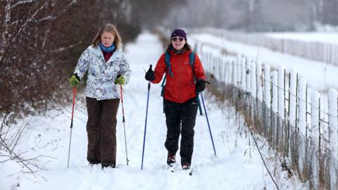 Two people use skis and walking poles to trek through a snowy path next to a snow-covered fence with trees in the background, in Banchory, Scotland on Friday.